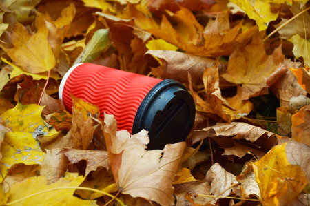 disposable paper cup lies on yellow maple leaves. garbage on autumn leaves. warming up while walking through the autumn park. warming drink: tea, coffee or cappuccinoの写真素材