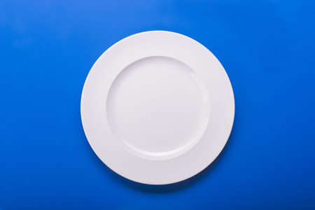 Empty white ceramic plate on blue background. top viewの写真素材