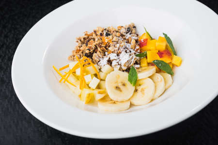 delicious granola with fruits, banana in white plate. healthy breakfast on black tableの写真素材
