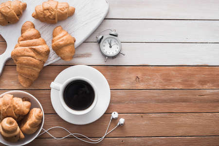 white cup of coffee with croissant, alarm clock and music headphones on a wood table. top view. cheerful start to the dayの写真素材