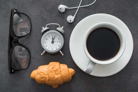 coffee cup with croissant and alarm clock, black glasses, white headphones. light snack during work. concept of business lunch. top viewの写真素材