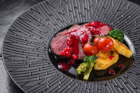 Beautiful serving of steak in the restaurant. Veal of medium roast with cranberry sauce, tomatoes and broccoli in a black plate.の写真素材