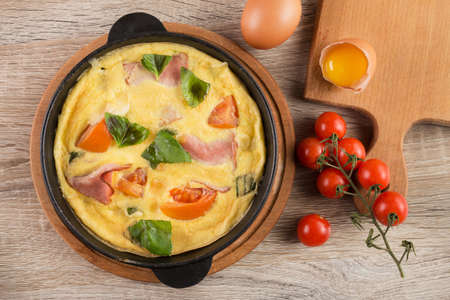 Hot omelet with bacon, tomato and basil on a frying metal pan stands on a wooden table. hearty breakfast. top viewの写真素材