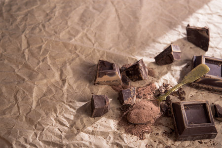Large pieces of dark chocolate and cocoa powder in vintage spoon, craft paper on a background. top...