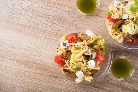 Plastic take away container with fresh salad ( mussels, feta cheese, pasta, tomato) on a wooden table.  Top view.の写真素材