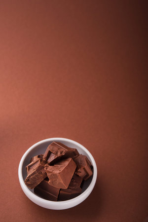 Pieces of chocolate in a white bowl on brown color background