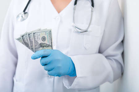 Doctor in clinic with a bundle of dollar bills. Corruption and bribery in medicine. Paid treatment.の写真素材