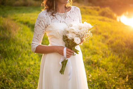 Bride in a dress holds a wedding bouquet of white roses, is in the park at sunset.