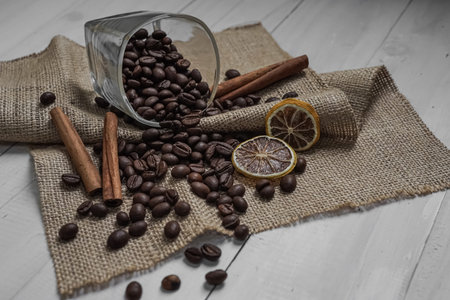 Roasted coffee beans scattering from glass on old rustic burlap with cinnamon sticks and dried...