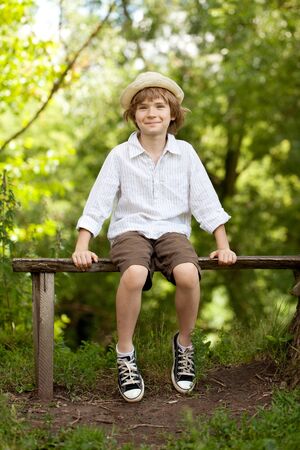 Boy in the hat and shorts sitting on a bench and smilingの写真素材