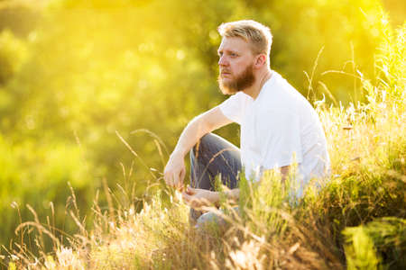 Blonde bearded young man in white t-shirt sits on the grass on a summer dayの写真素材