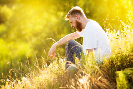 Blonde bearded young man in a T-shirt sits on the grass on a summer dayの写真素材