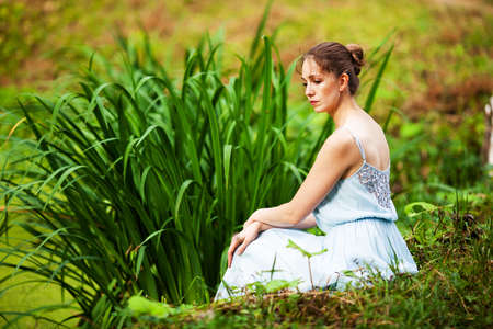 Young woman in a blue dress sits on the grass by the pondの写真素材