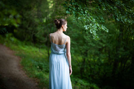 Young woman in a blue dress stands with her back to us in the parkの写真素材