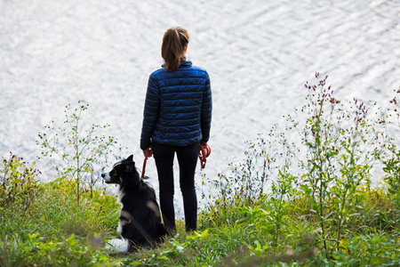 Slender young woman stands on the shore of the lake with her dogの写真素材