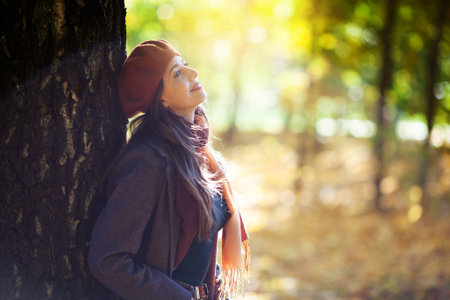 Girl in an orange beret stands next to a tree on a sunny autumn dayの写真素材