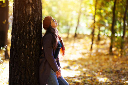 Girl in a beret stands leaning against a tree on a sunny autumn dayの写真素材