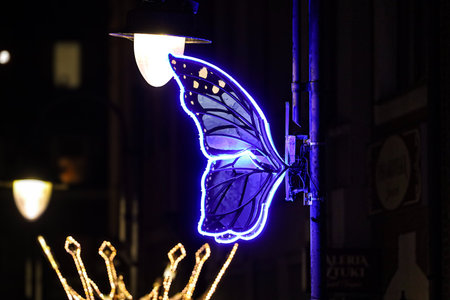 A striking blue neon butterfly wing art installation is mounted on a pole, illuminated against the dark night sky with a warm street lamp nearbyの写真素材
