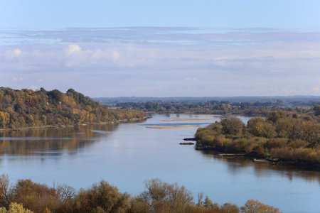 A wide view of the Vistula River with calm water reflecting the sky. Autumn foliage in yellow and...