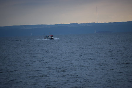 The photo shows a small boat sailing on the sea, leaving a trail behind. Shoreline with hills and...