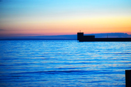 The photo shows a sunset over the sea. The sky is bright orange and yellow with silhouettes of buildings and trees on the horizon. The water reflects the sky colors creating a harmonious landscapeの写真素材