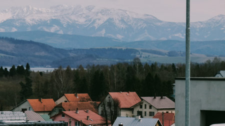 Two houses on green fields, forest behind, and snow-covered Tatras mountains on horizon under light sky.の写真素材