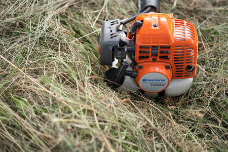 Minsk, Belarus - 02/08/2020 Petrol scythe trimmer brushcutter Husqvarna 143R-II lies on the mown hay after mowing tall grass in the garden of a country house.のeditorial素材