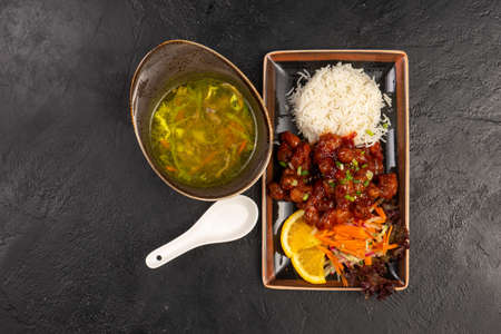 Lunch menu of vegetable soup and hot meat dish with rice and fresh vegetables as a side dish. Two Asian dishes in unusual traditional dishes on a black stone table.の写真素材