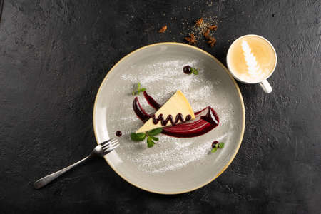 Classic dessert with coffee - cheesecake with berry sauce, garnished with mint leaves and powdered sugar on a round ceramic plate and a cup of cappuccino on a black stone kitchen table.の写真素材