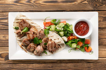 Pork shish kebab in lavash with ketchup and vegetable salad of cucumbers, tomatoes and bell peppers in a white ceramic plateの写真素材