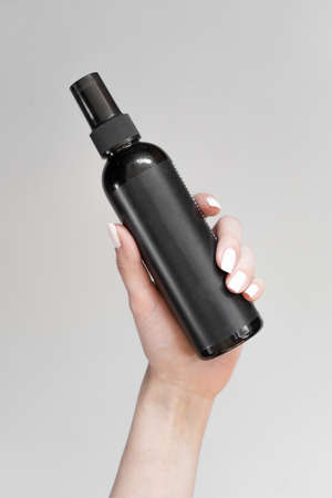 A black bottle of cosmetic spray with an empty label in a woman's hand on a light backgroundの写真素材