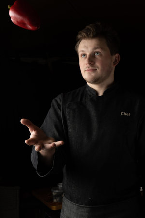 A cook in a black jacket throws up and catches a red sweet pepper with his hand