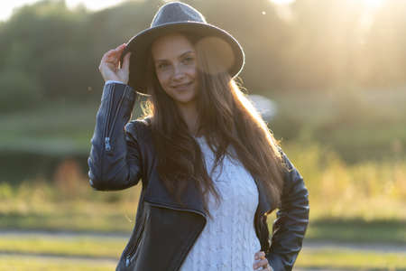 A young, beautiful long-haired woman in a hat smiles, looks at the camera in the park in the bright glow of sunset raysの写真素材
