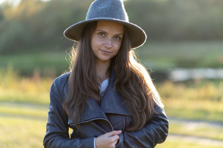 A young, beautiful long-haired woman in a hat smiles, looks at the camera in the park in the bright glow of sunset raysの写真素材