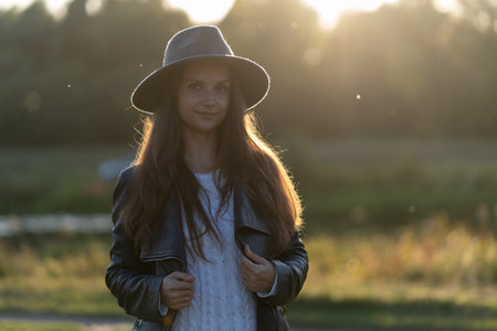 A young, beautiful long-haired woman in a hat smiles, looks at the camera in the park in the bright glow of sunset raysの写真素材