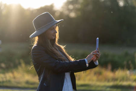 A young, beautiful woman in a hat smiles, takes a selfie and takes a video in the park on her smartphone in the bright radiance of the rays of the sunset raysの写真素材