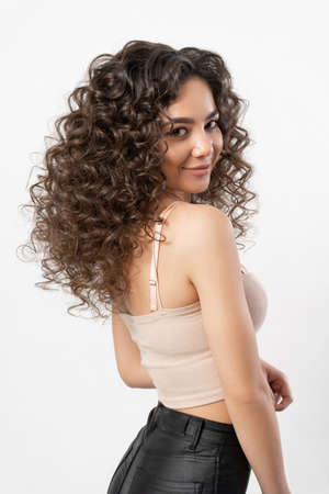 A beautiful young female model with curly long dark hair curled on a curling iron smiles and poses for a portrait on a white background.の写真素材
