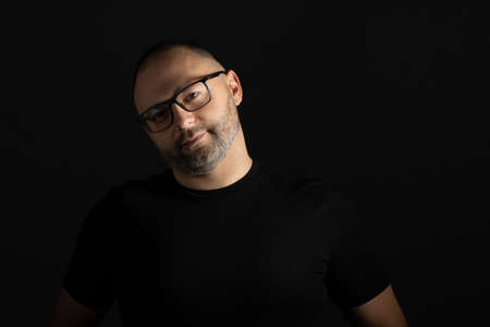 A young man in a black T-shirt with a short haircut and black-rimmed glasses poses against a dark background carefully and aggressively looking at the cameraの写真素材