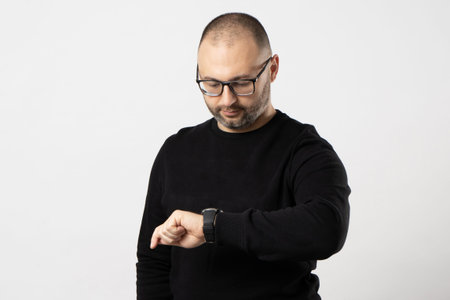 Portrait of a man with black-rimmed glasses, short haircut and a black jacket looking at a clock on...