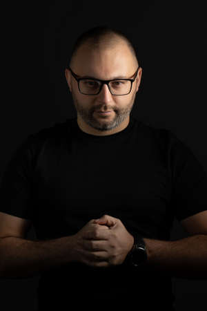A young man in a black T-shirt with a short haircut and black-rimmed glasses poses against a dark background carefully and aggressively looking at the cameraの写真素材