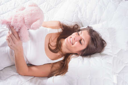 Beautiful young woman lying in bed and holding teddy bear while she smilingの写真素材