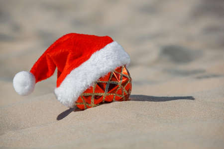 red Santa Claus hat wearing on Christmas ball lying on the beach, on the sand. New Year's holidays in the ocean or sea.の写真素材
