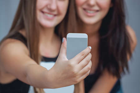 Two young, attractive, smiling woman holding a cell phone and make a selfie.の写真素材
