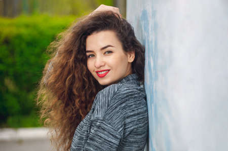 Portrait of an attractive young woman with curly hair standing with her back to the wall and smiling.の写真素材
