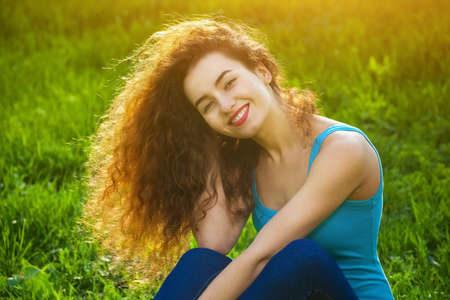 Attractive, young girl with curly hair sitting on the green grass on the lawn and smiling at the photographer on the background of sunset.の写真素材