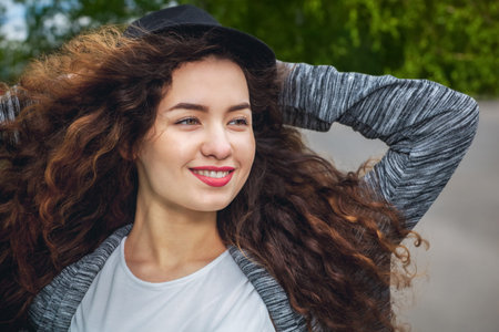 Attractive, young girl with curly hair and a hat on a background of green trees on the lawn and...