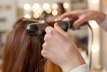 Hairdresser makes hairstyle girl with long red hair in a beauty salon. Create curls with curling irons. Professional hair care.の写真素材
