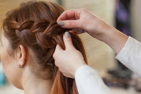 Beautiful, red-haired girl with long hair, hairdresser weaves a French braid, in a beauty salon. Professional hair care and creating hairstyles.の写真素材