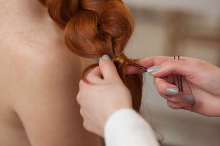 Beautiful, red-haired girl with long hair, hairdresser weaves a French braid, in a beauty salon. Professional hair care and creating hairstyles.の写真素材