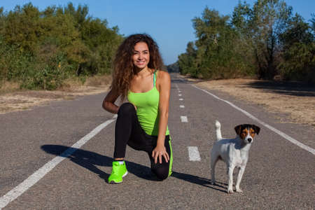 Attractive curly girl athlete doing morning exercises with her dog on the street. The concept of sports and healthy lifestyle.の写真素材